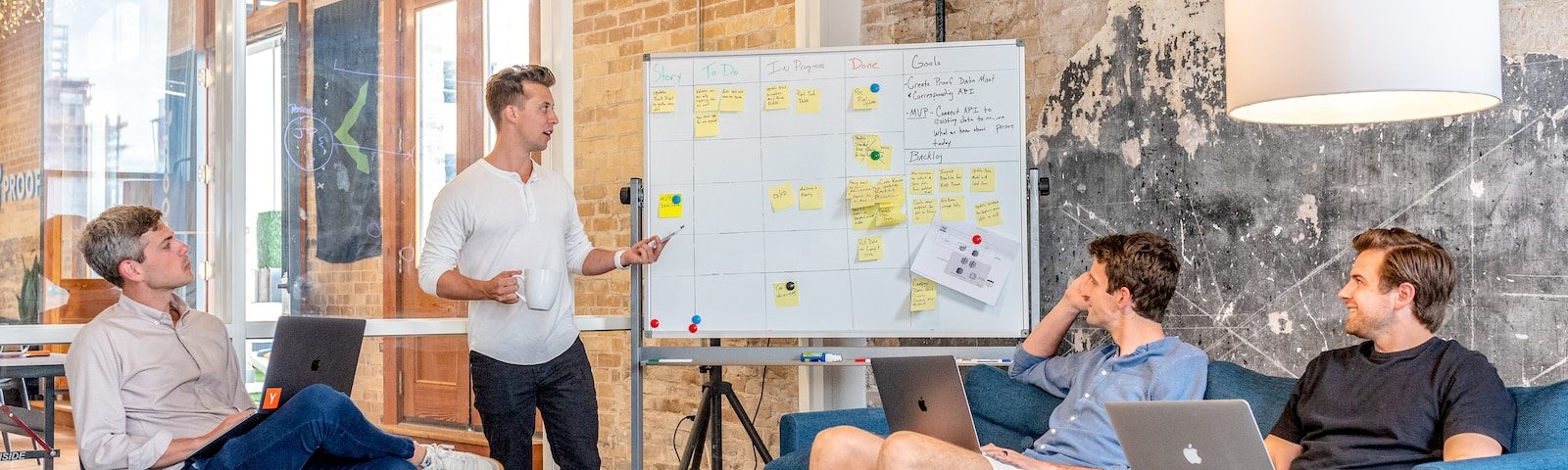 three men sitting while using laptops and watching man beside whiteboard